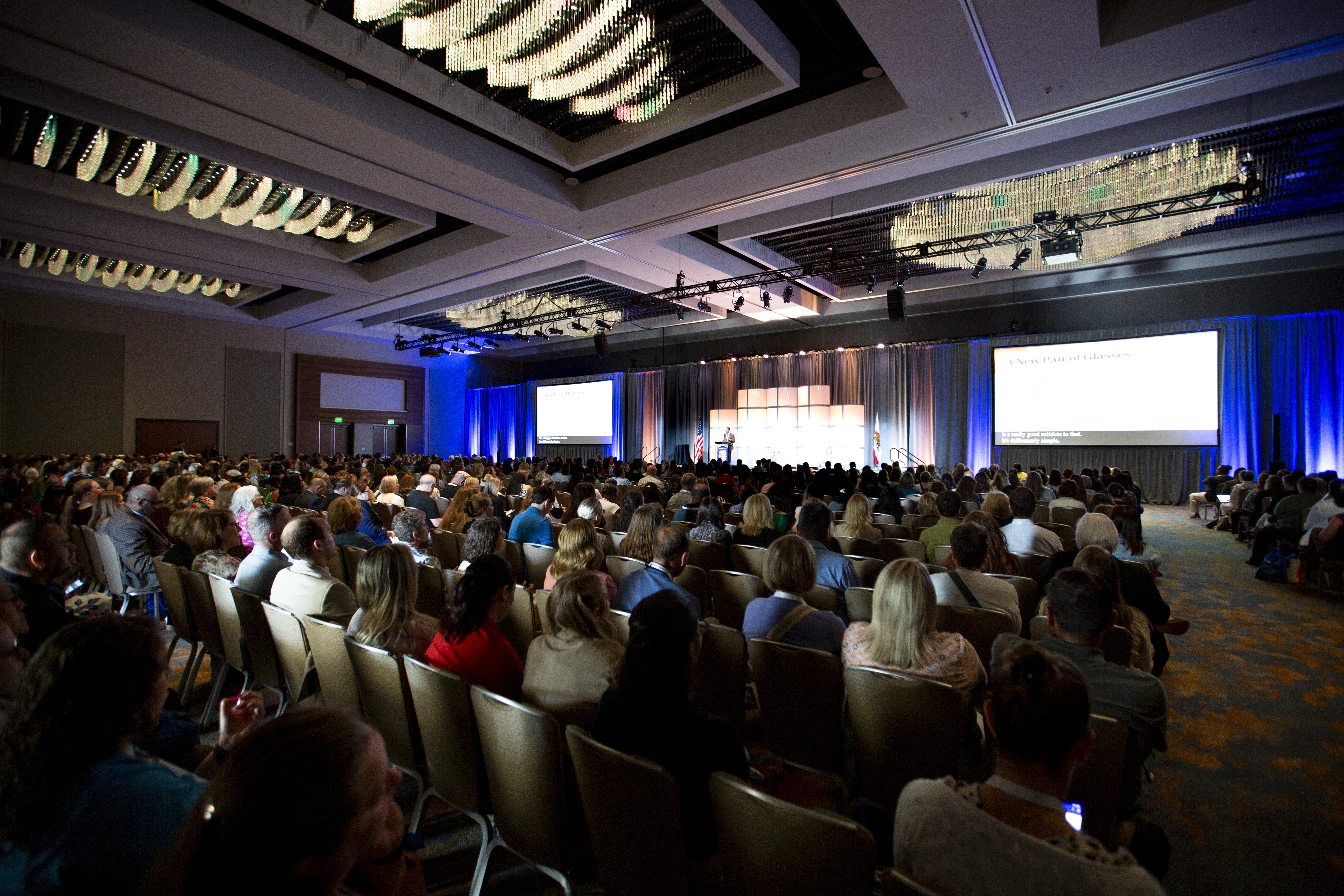 An audience seated in a large ballroom listen to a speaker on stage at the USAging 51st Annual Conference and Tradeshow. The room is filled with hundreds of attendees, with presentation screens and blue lighting at the front of the stage.