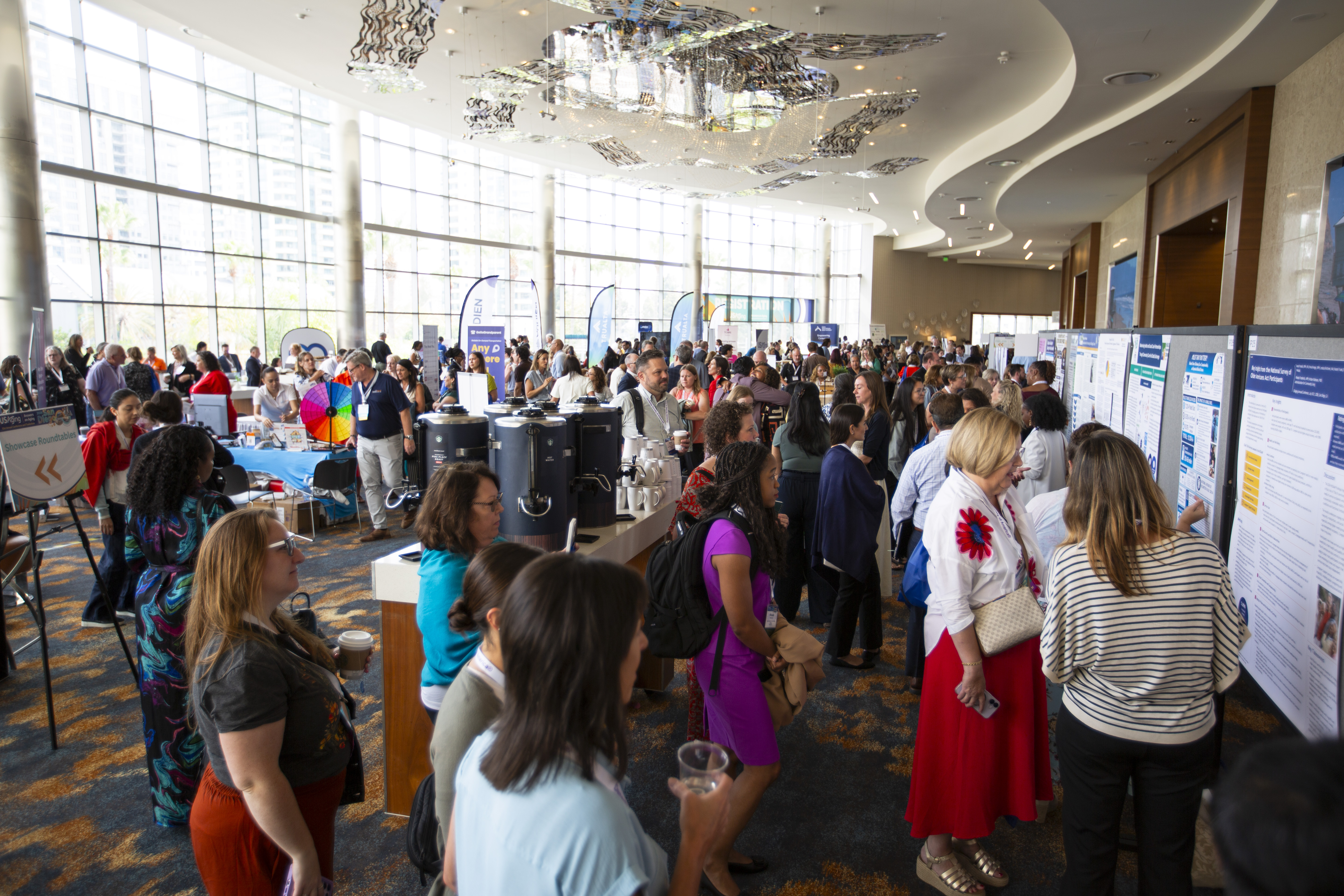 Attendees in the Discovery Showcase. The room is filled with people. Some are looking at the Posters in the Poster Session and talking with the poster presenters. Others are at tables on the opposite side of the room from the posters, speaking with exhibitors at tables who are demonstrating their products. Food stations are in the center of the room, where attendees pick up coffee and snacks.