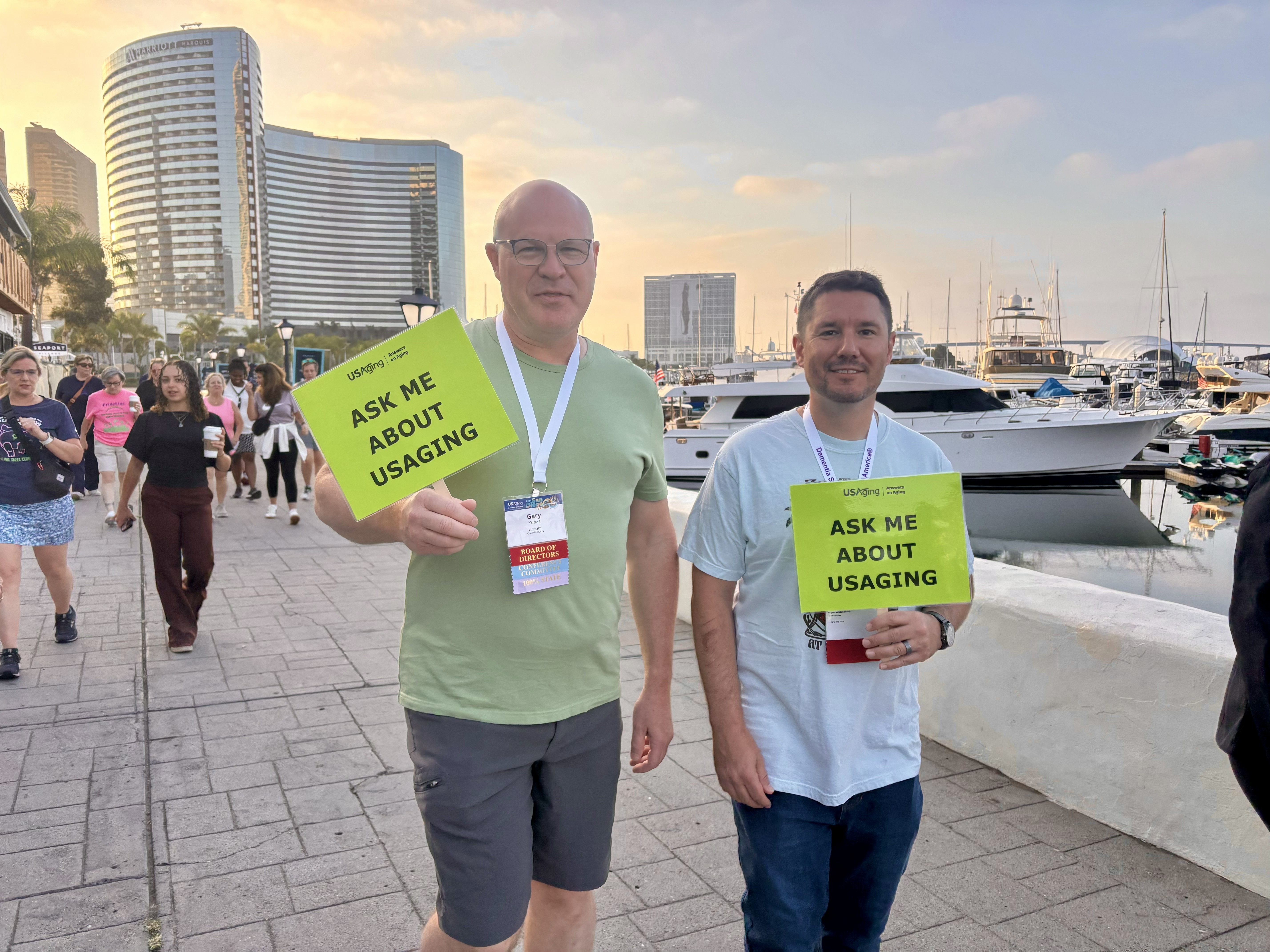 USAging Board members Gary Yuhas and Justin Lees participate in the 2026 Early Bird Walk along the San Diego Embarcadero and the bay. They are dressed in Tshirts and shorts and are holding signs that read “Ask Me About USAging.” Behind them, the sky is pink from the sunrise and boats are docked along the way.